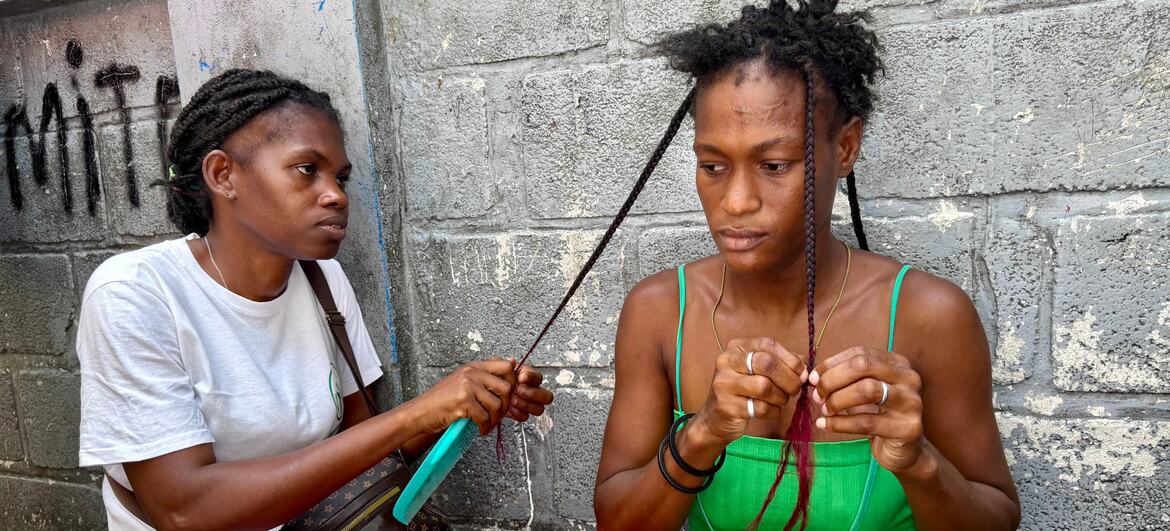 Two women are sitting against a concrete wall, braiding hair.