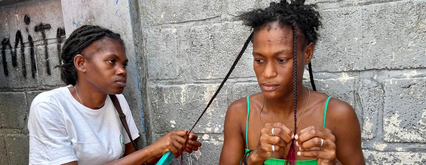 Deux femmes sont assises contre un mur en béton, en train de tresser des cheveux.