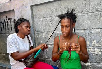 Deux femmes sont assises contre un mur en béton, en train de tresser des cheveux.