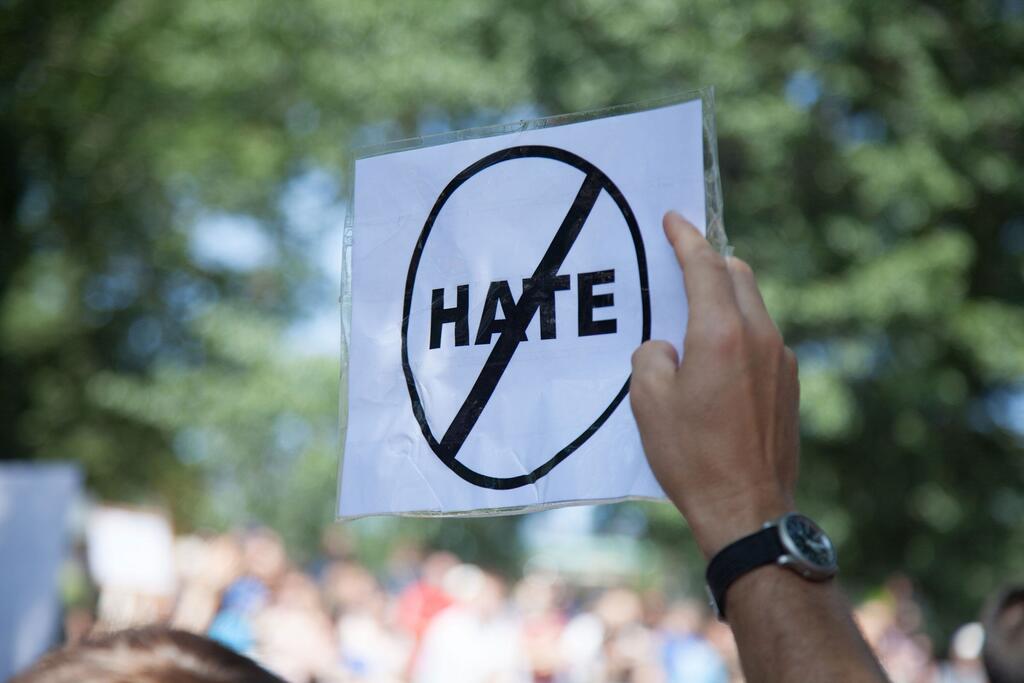 Lors d'une manifestation, une personne brandit une pancarte où l'on peut lire « Non à la haine », le mot « haine » étant barré d'un cercle et d'une barre oblique. En arrière-plan, on aperçoit une foule et des arbres.
