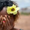 Une femme à la peau mate, une fleur jaune dans les cheveux, se cache le visage avec ses mains. Ses ongles sont vernis en rose. L'arrière-plan est un paysage extérieur flou.