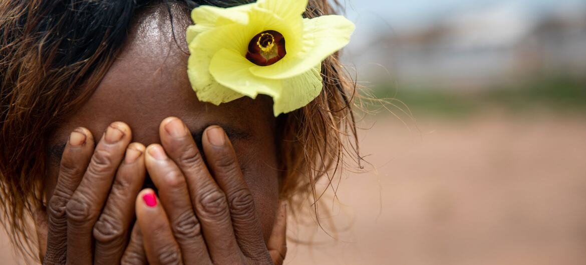 Una mujer de piel morena con una flor amarilla en el cabello se cubre el rostro con las manos. Tiene las uñas pintadas de rosa. El fondo es una escena exterior borrosa.