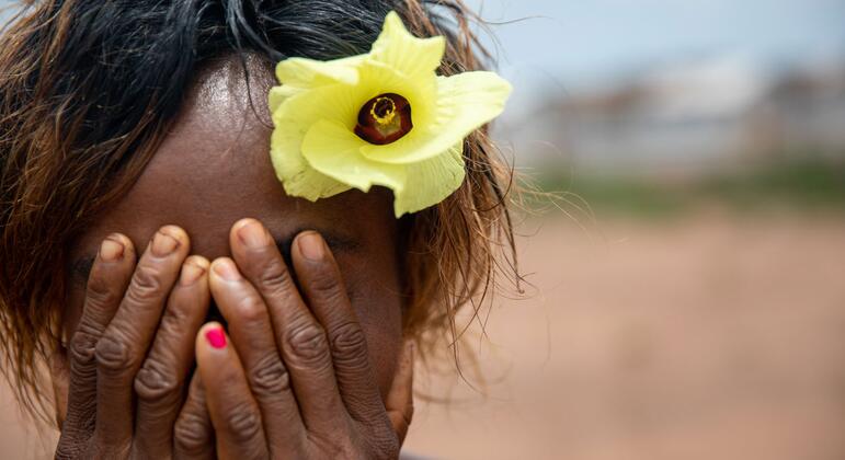 Une femme à la peau mate, une fleur jaune dans les cheveux, se cache le visage avec ses mains. Ses ongles sont vernis en rose. L'arrière-plan est un paysage extérieur flou.