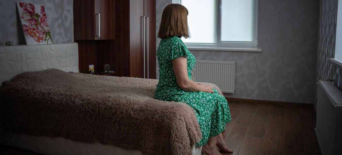 A woman in a green dress sits on a bed in a bedroom, looking out the window.