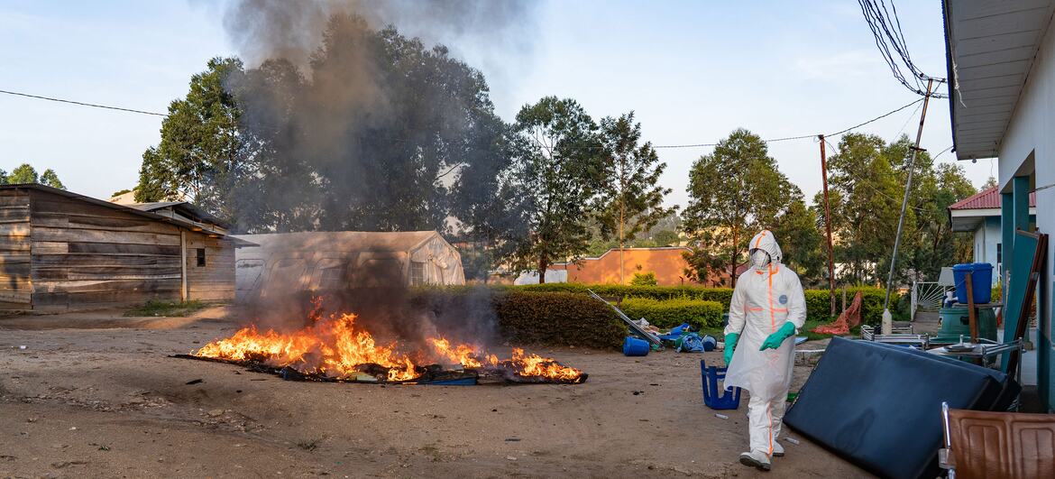 Un agent de santé, entièrement équipé d'une protection, se tient près d'un grand bûcher à ciel ouvert où brûle un corps dans le cadre de la réponse de l'OMS à une épidémie d'Ebola en République démocratique du Congo.