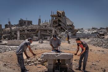 Three men work with a piece of metal on a makeshift concrete table amidst the rubble of a destroyed building in Gaza.