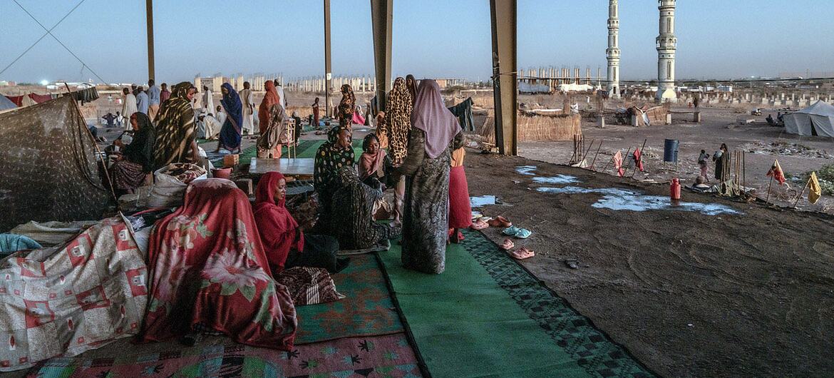 IDP settlement in Gedaref, Sudan, showing displaced people in a former bus station, with a mosque in the background.