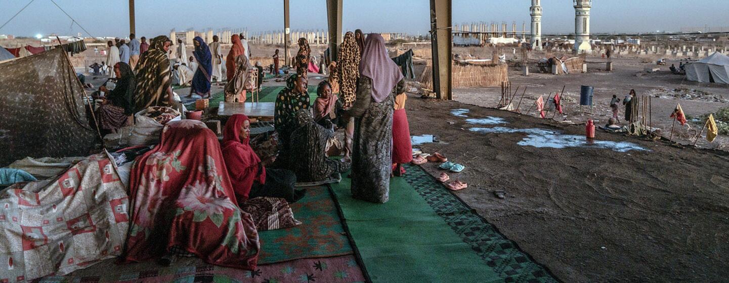 Camp de déplacés à Gedaref, au Soudan, montrant des personnes déplacées dans une ancienne gare routière, avec une mosquée en arrière-plan.