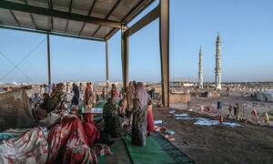 Camp de déplacés à Gedaref, au Soudan, montrant des personnes déplacées dans une ancienne gare routière, avec une mosquée en arrière-plan.