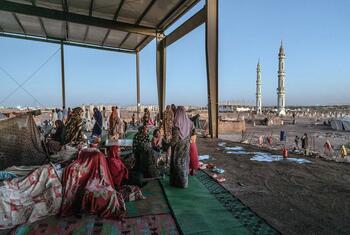 IDP settlement in Gedaref, Sudan, showing displaced people in a former bus station, with a mosque in the background.