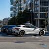 A white Waymo self-driving car, a modified Tesla Model Y, waits at an intersection in a modern urban setting on a sunny day.