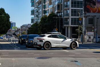 A white Waymo self-driving car, a modified Tesla Model Y, waits at an intersection in a modern urban setting on a sunny day.