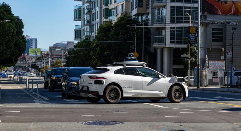 Un coche autónomo blanco de Waymo, un Tesla Model Y modificado, espera en una intersección en un entorno urbano moderno en un día soleado.