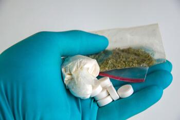 一只戴着手套的手拿着一些违禁品样品，包括药丸、一袋白色粉末和一小袋绿色草药。