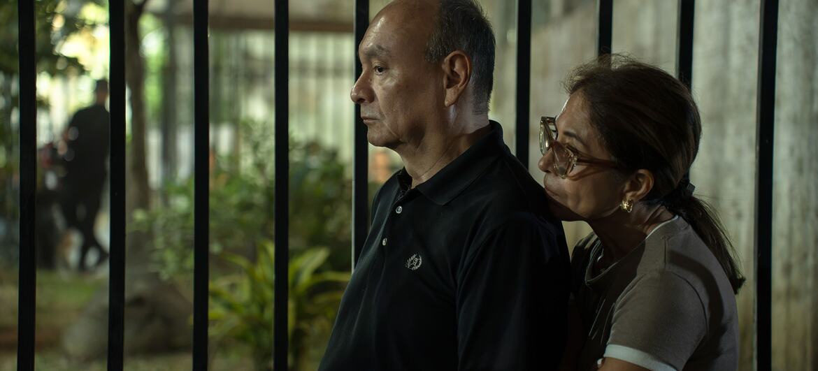 Un hombre y una mujer venezolanos permanecen de pie uno al lado del otro detrás de una valla por la noche, mirando hacia otro lado con expresiones de dolor y preocupación tras el terremoto de 2026.