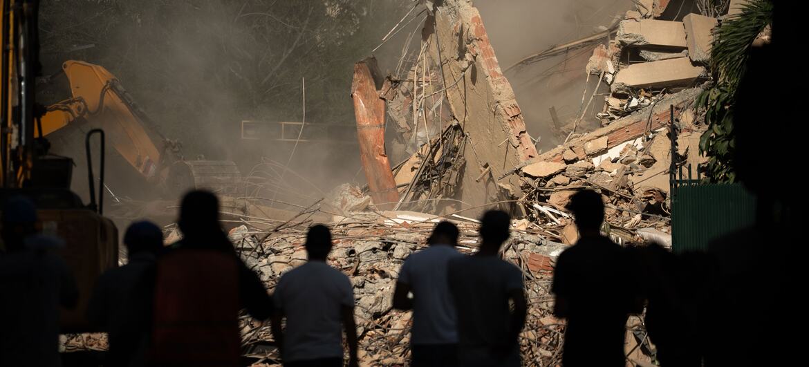 Personal de emergencia y maquinaria pesada en el lugar del derrumbe de un edificio, con polvo levantándose de los escombros.