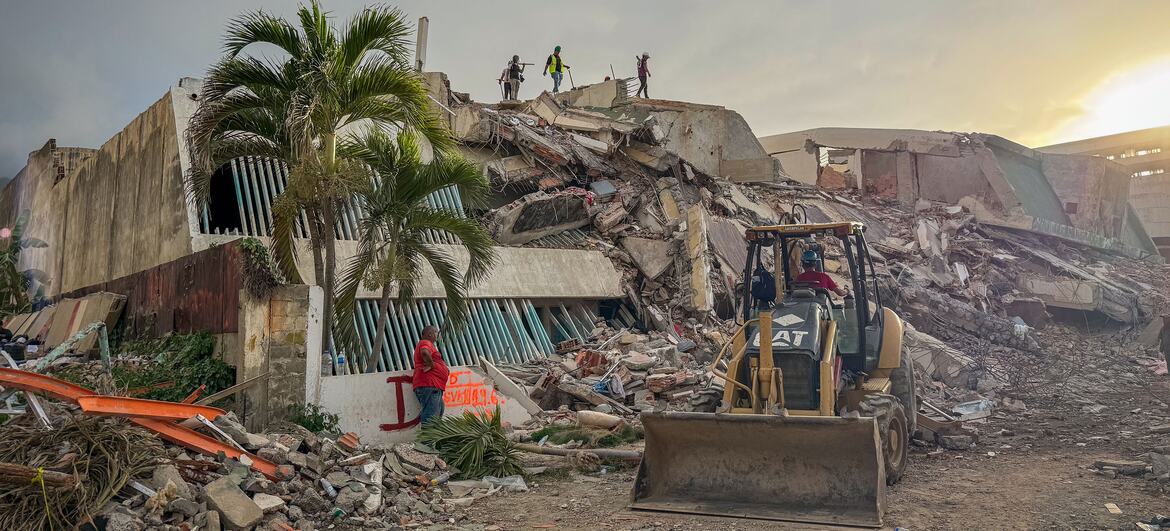 Photo by UNOCHA/Veronique Durroux showing search and rescue operations near the earthquake ground zero in La Guaira, Venezuela, on June 29, 2026. A CAT backhoe loader is positioned in the foreground amidst rubble, with several rescue workers visible on top of the collapsed building structure.