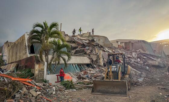 Photo by UNOCHA/Veronique Durroux showing search and rescue operations near the earthquake ground zero in La Guaira, Venezuela, on June 29, 2026. A CAT backhoe loader is positioned in the foreground amidst rubble, with several rescue workers visible on top of the collapsed building structure.