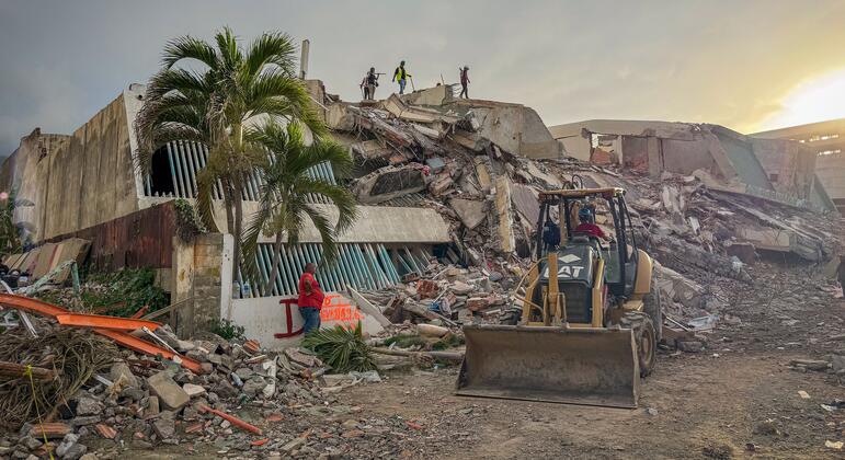 Photo by UNOCHA/Veronique Durroux showing search and rescue operations near the earthquake ground zero in La Guaira, Venezuela, on June 29, 2026. A CAT backhoe loader is positioned in the foreground amidst rubble, with several rescue workers visible on top of the collapsed building structure.