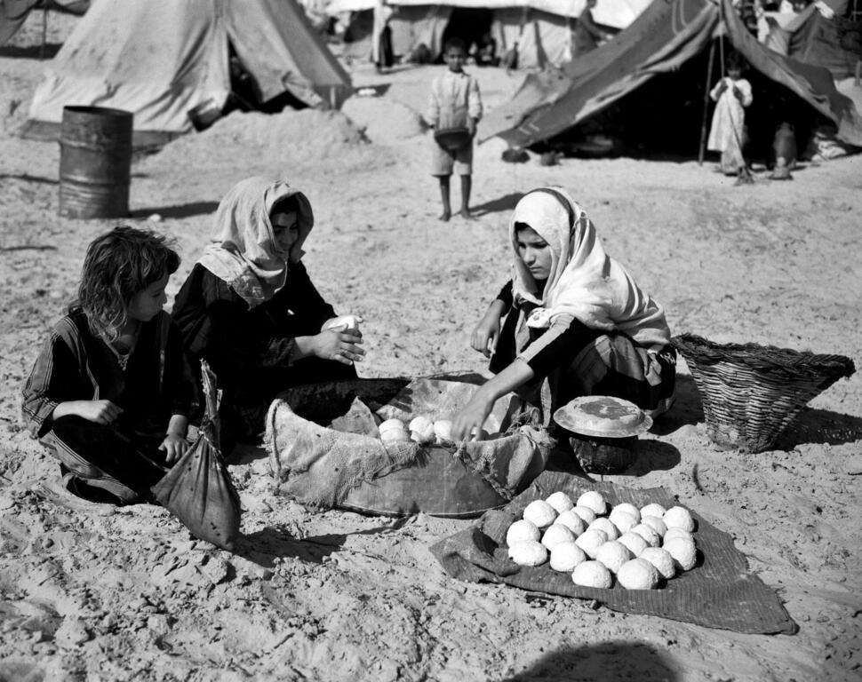 Palestinian women and a child prepare food in a refugee camp with tents in the background.