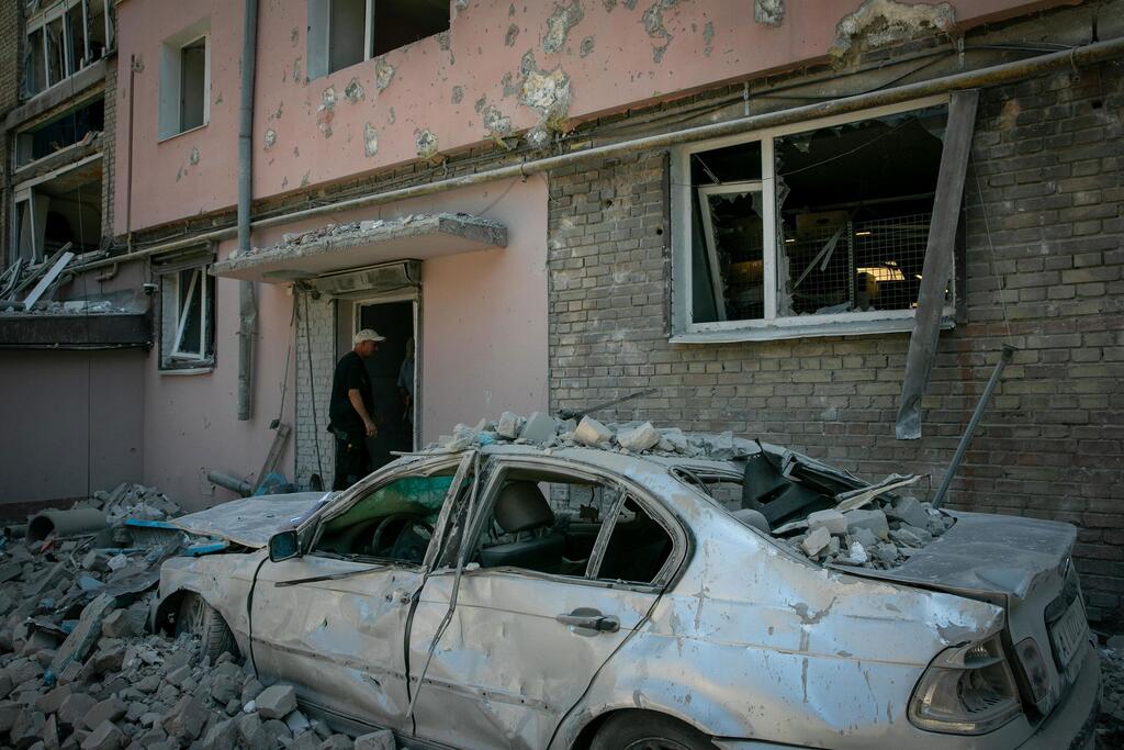 À Kyiv, en Ukraine, une voiture argentée endommagée, recouverte de gravats de béton, est garée devant un immeuble en briques roses aux fenêtres brisées. Un homme se tient sur le seuil de l'immeuble.