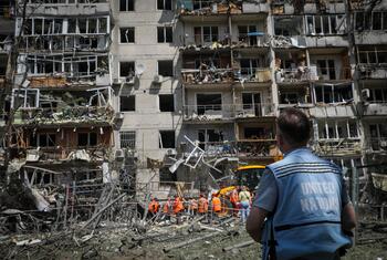 Au premier plan, un homme portant un gilet des Nations Unies observe un immeuble d'habitation fortement endommagé à Kyiv, en Ukraine, où des équipes de recherche et de sauvetage travaillent parmi les décombres.