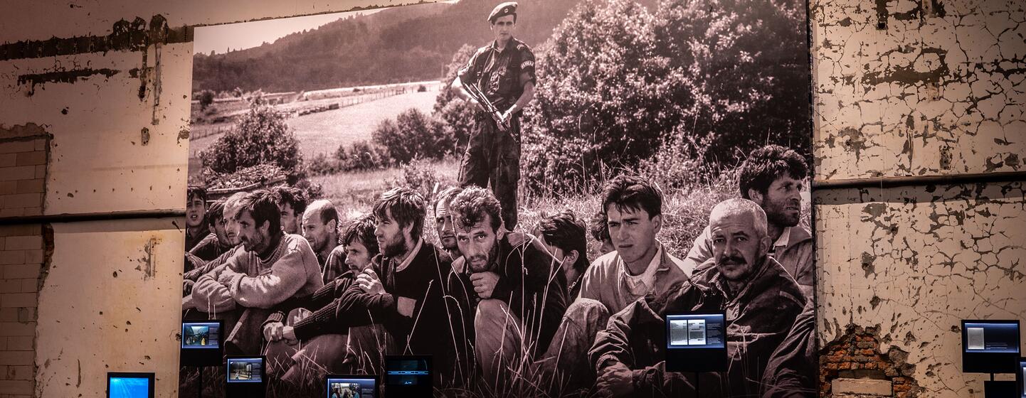 A large historical photograph displayed at the Srebrenica memorial, showing survivors and victims of the 1995 massacre. The exhibit includes informational text panels and video screens.