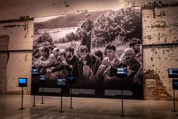 A large historical photograph displayed at the Srebrenica memorial, showing survivors and victims of the 1995 massacre. The exhibit includes informational text panels and video screens.