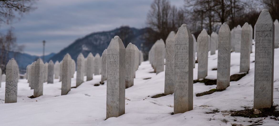Rows of white tombstones in a cemetery covered with snow, with mountains in the background.