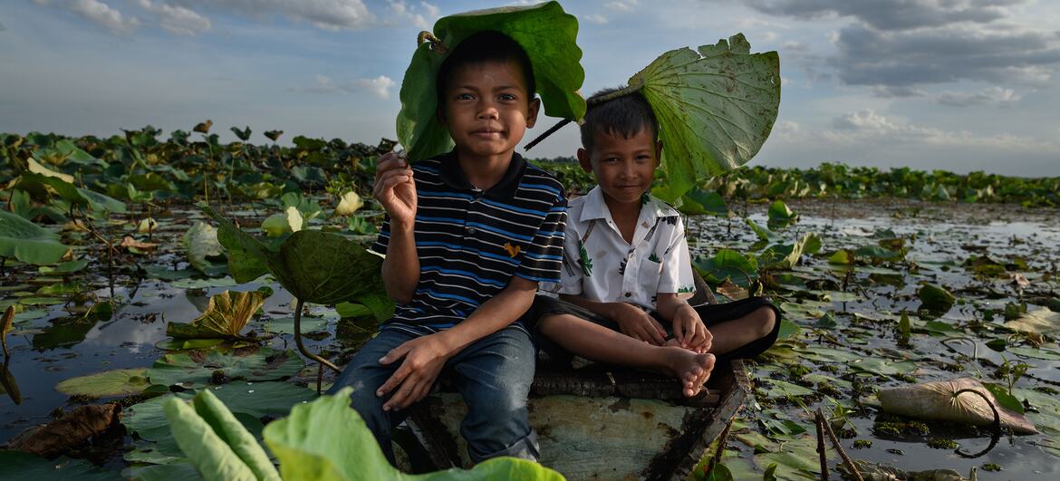 Two Thai children sit on a small wooden raft in a lotus pond, using large leaves as shade.