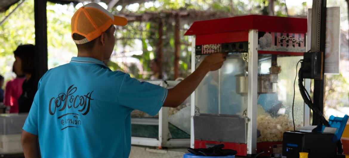 A man in a blue shirt and orange cap operates a popcorn machine at an outdoor street food stall.