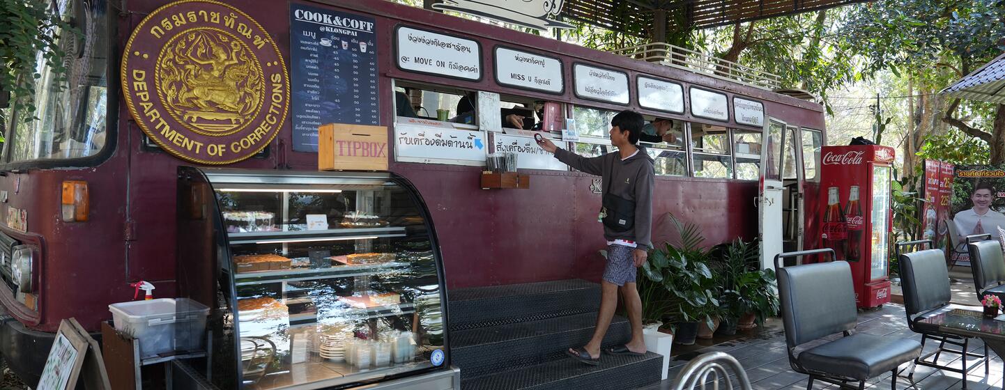 A man orders food at a mobile coffee shop built into a red bus, with a food display case in front. The bus has a 'Department of Corrections' seal and a 'Cook & Coffee' sign, located on a wooden deck under a roof with trees in the background.
