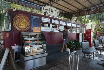 A man orders food at a mobile coffee shop built into a red bus, with a food display case in front. The bus has a 'Department of Corrections' seal and a 'Cook & Coffee' sign, located on a wooden deck under a roof with trees in the background.