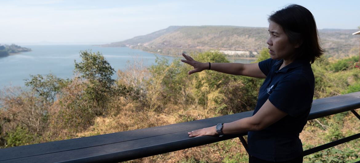 An Asian woman in a dark polo shirt points towards a lake and hills from a viewing platform in Thailand.