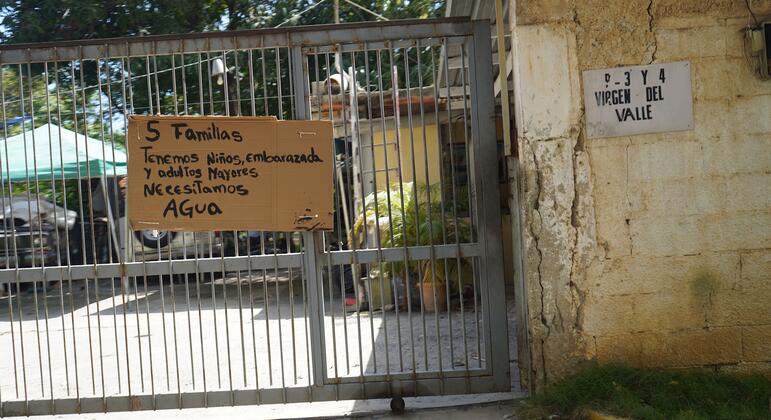 A handwritten sign hangs on the front gate of a damaged house in La Guaira, Venezuela, reading: '5 families, we have children, a pregnant woman, and elderly people, we need water'.