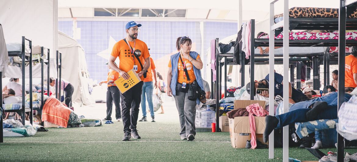 Los trabajadores humanitarios del UNFPA , vestidos con camisetas naranjas, se encuentran en un campamento de socorro ubicado dentro de una gran estructura de tiendas de campaña con literas provisionales para personas desplazadas.