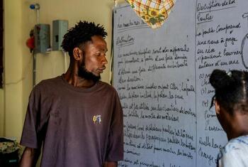 A Haitian man, Edens Désir, stands in an improvised school shelter in Hinche, Haiti, next to a whiteboard with French writing, while a child looks on.