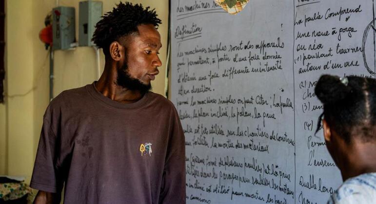 A Haitian man, Edens Désir, stands in an improvised school shelter in Hinche, Haiti, next to a whiteboard with French writing, while a child looks on.