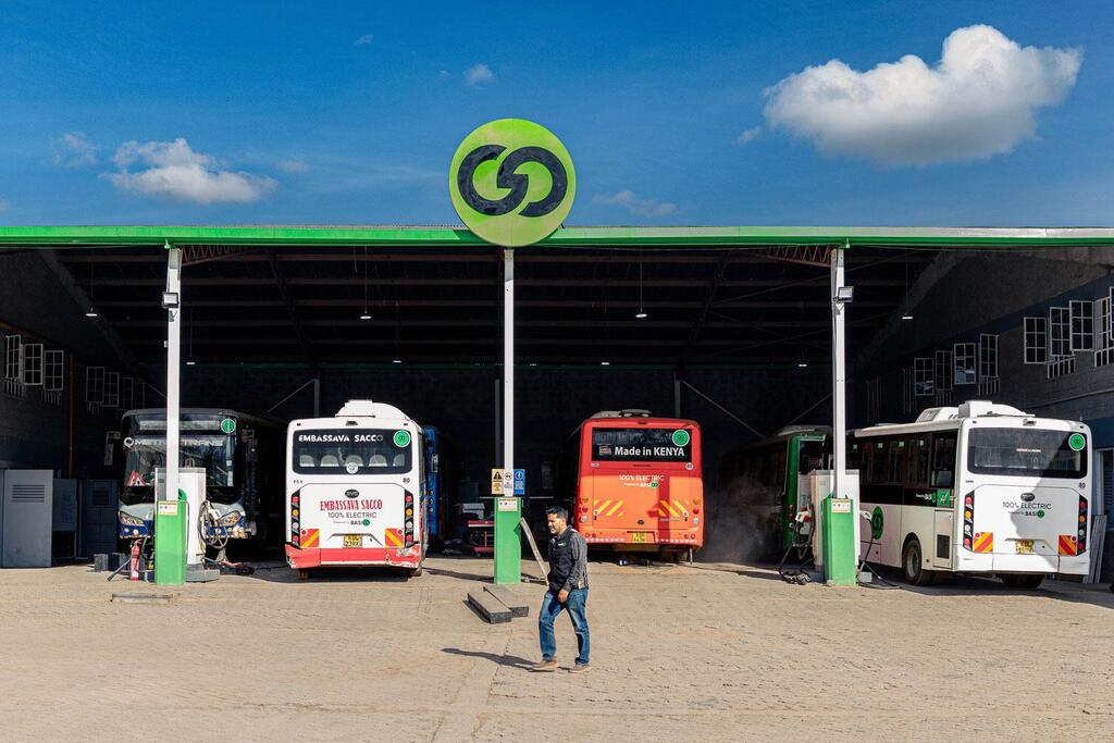 Plusieurs bus électriques à une station de recharge sous un auvent vert, avec un homme qui marche au premier plan.