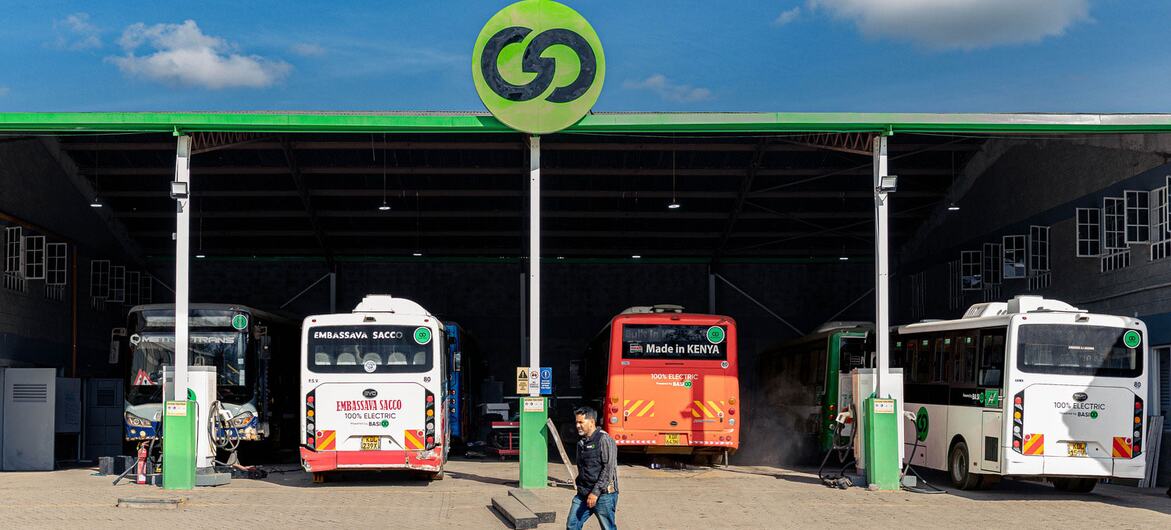 Several electric buses at a charging station under a green canopy, with a man walking in the foreground.