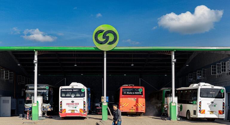 Plusieurs bus électriques à une station de recharge sous un auvent vert, avec un homme qui marche au premier plan.