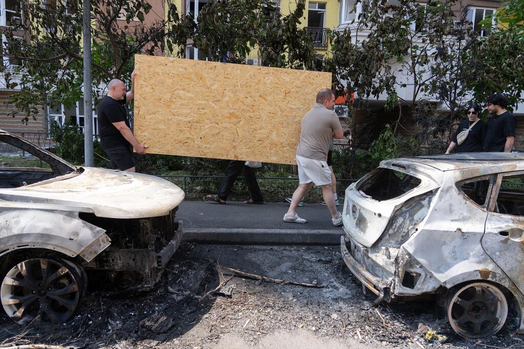 Deux hommes transportent une grande plaque de contreplaqué devant des voitures calcinées dans une rue de Kyiv, en Ukraine.
