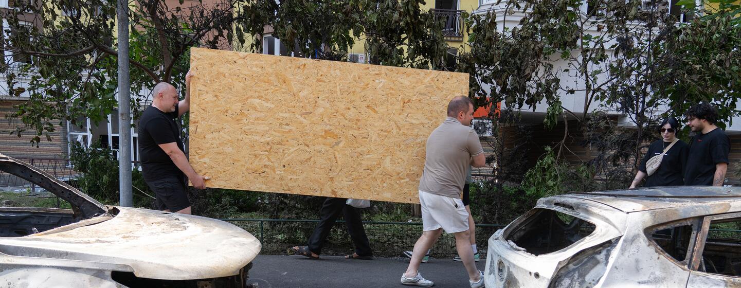 Two men carry a large sheet of plywood past burnt-out cars in a street in Kyiv, Ukraine.