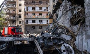 A fire truck is present amidst the wreckage of a destroyed car and a damaged apartment building in Kyiv, Ukraine.