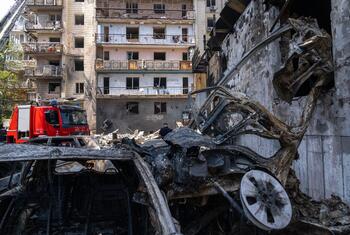 A fire truck is present amidst the wreckage of a destroyed car and a damaged apartment building in Kyiv, Ukraine.