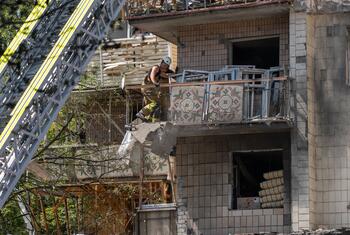 A firefighter in protective gear stands on a damaged balcony of a destroyed building in Kyiv, Ukraine, while a ladder truck is positioned nearby.
