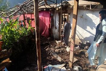 Men in traditional clothing stand in the yard of a rural homestead with chickens on a muddy floor.