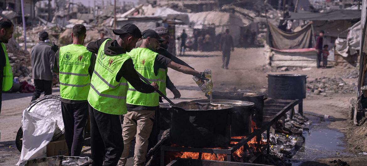 WFP aid workers in green vests prepare and distribute hot meals to families in the Jabalyiah area of Gaza, with a backdrop of destroyed buildings.