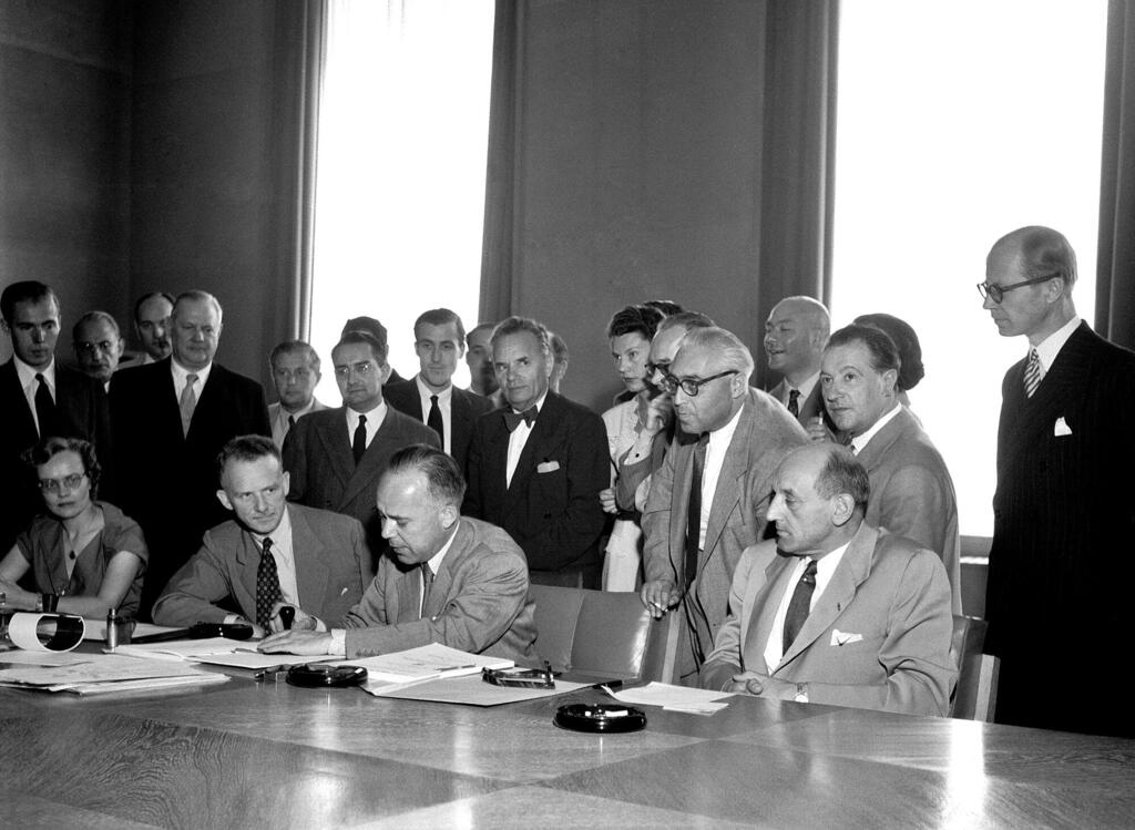 Maafisa katika Palais des Nations huko Geneva wanasaini Mkataba Unaohusiana na Hali ya Wakimbizi mwaka wa 1951. Walioketi mezani ni, kutoka kushoto kwenda kulia, Margaret Kitchen, John Humphrey, Knud Larsen, na Dkt. GJ Van Heuven Goedhart, huku wajumbe na waangalizi wengine wakiwa wamesimama nyuma yao.