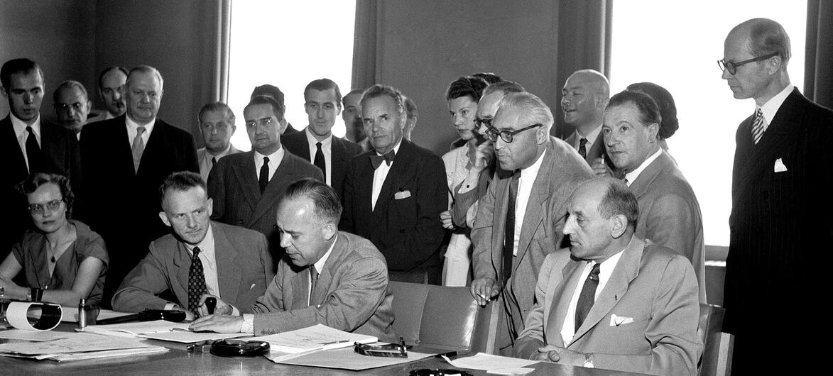 En 1951, des officiels du Palais des Nations à Genève signent la Convention relative au statut des réfugiés. Assis à la table, de gauche à droite : Margaret Kitchen, John Humphrey, Knud Larsen et le Dr GJ Van Heuven Goedhart, avec d’autres délégués et observateurs debout derrière eux.